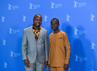 William Kamkwamba (esq.) e Maxwell Simba, ator do filme “O menino que descobriu o vento”, no Festival Internacional de Cinema de Berlim, em 12 de fevereiro de 2019. O filme se baseia na autobiografia de mesmo nome de William Kamkwamba, o mecânico nascido no Malawi que se tornou um herói em seu país natal ao construir um moinho de vento feito de eucalipto, peças de bicicleta e material do ferro-velho local, a fim de abastecer sua casa com eletricidade. Mais tarde, ele construiu uma bomba de água movida a energia solar, que forneceu água potável para sua aldeia pela primeira vez. Como sua família não podia pagar as taxas escolares, Kamkwamba teve que deixar a escola. No entanto, ele continuou sua educação na biblioteca da aldeia, onde descobriu o livro “Usando energia”. Foi nele que Kamkwamba viu a imagem e a explicação do funcionamento de um moinho de vento.