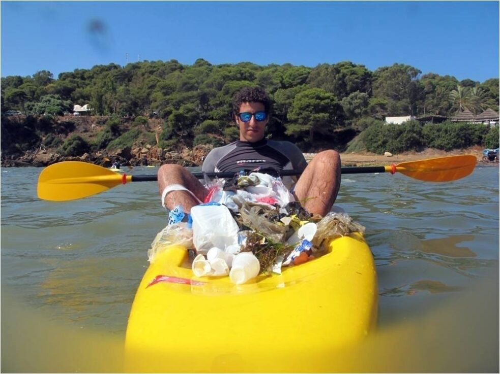 Personne assise dans un kayak rempli de déchets ramassés, face à la caméra.