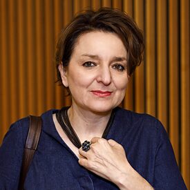 Square image of Eva Illouz against a brown backdrop; she wears short dark hair and holds a glas in her left hand