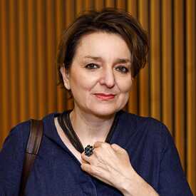 Quadratisches Bild von Eva Illouz vor bräunlichem Hintergrund; sie hat schwarze, kurze Haare und hält ein Glas in der linken Hand