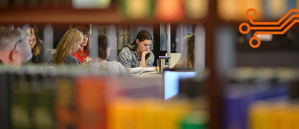 Blick durch ein Bücherregal: Teilnehmende eines Workshops sitzen an einem Tisch.