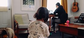 Ett litet barn sitter i ett rum, i bakgrunden sitter hans mor vid skrivbordet och arbetar.