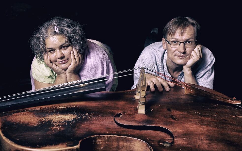 Zwei Personen liegen hinter einem Kontrabass und lächeln in die Kamera