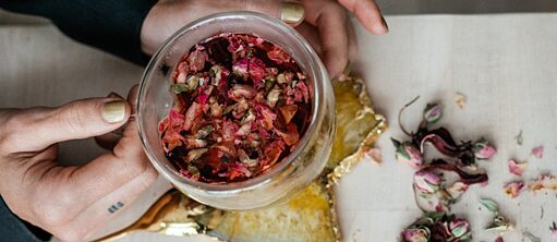 Two hands hold a cup of pink herbal tea at a white table, with flowers and herbs scattered on the table