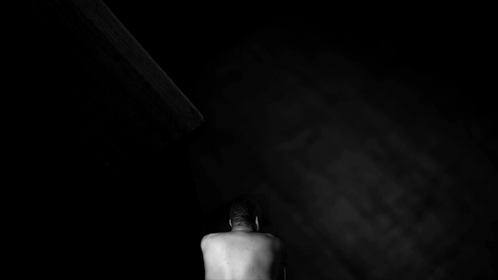 Colonial history – In this screenshot, Aurêlio Valente Langa is sitting in a dark room. His naked back is turned toward the camera.