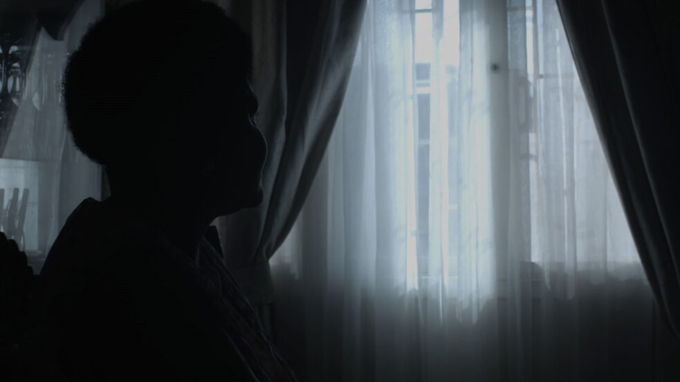 Colonial history – This screenshot depicts Isabel Langa who is sitting in a darkened room and looking out the window.