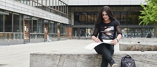 Eine junge Frau sitzt vor einem öffentlichen Gebäude. Sie öffnet eine Mappe auf ihrem Schoß, neben ihr ein Rucksack auf dem Boden.
