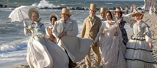 Mehrere Personen in historischen weißen und beigen Kostümen um 1900 gehen an einem Strand spazieren.