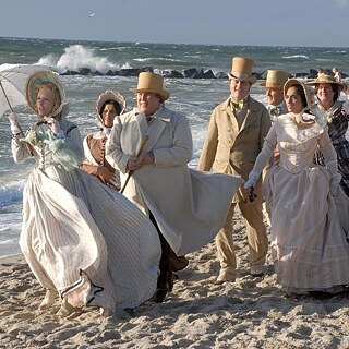 Mehrere Personen in historischen weißen und beigen Kostümen um 1900 gehen an einem Strand spazieren.