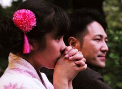Scène  du film 'Family Romance'. Deux visages de profils côte à côte : une jeune fille japonaise portant un pompon rose dans les cheveux porte les deux mains devant sa bouche, et regarde fixement devant elle. À ses côtés un jeune homme regarde dans la même direction