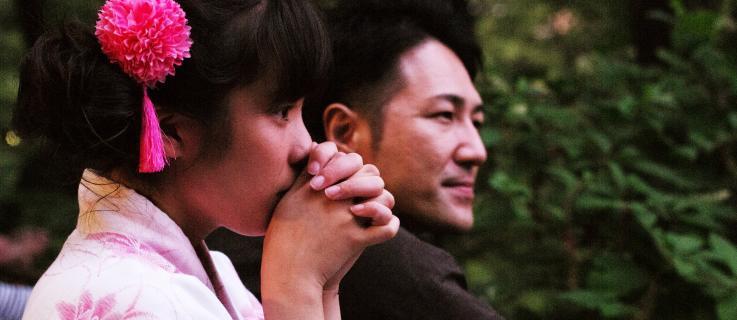 Scène  du film 'Family Romance'. Deux visages de profils côte à côte : une jeune fille japonaise portant un pompon rose dans les cheveux porte les deux mains devant sa bouche, et regarde fixement devant elle. À ses côtés un jeune homme regarde dans la même direction