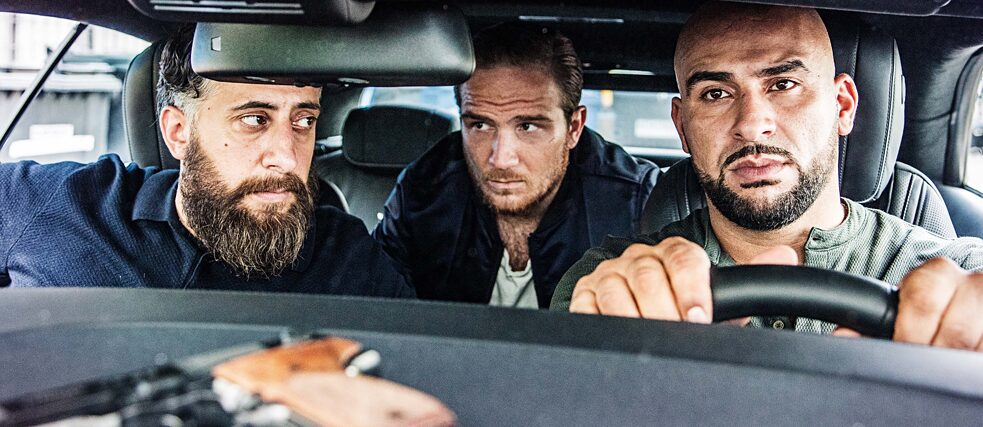 Image fixe de la série TNT "4 Blocks" Toni, Vince et Abbas vus à travers le pare-brise de leur voiture, une arme de poing posée sur l'étagère.