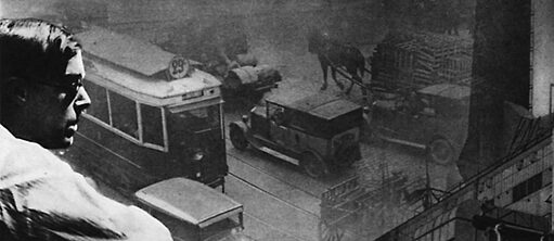 Un homme (à gauche) regarde une rue avec des voitures, un tram et une calèche.
