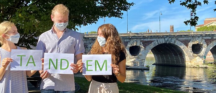 Zwei junge Frauen und ein junger Mann mit Masken und den Buchstaben TANDEM auf Wiese vor Brücke