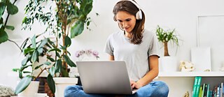 Eine Frau mit Kopfhörern sitzt zu Hause auf dem Boden und arbeitet lächelnd mit dem Laptop. Neben ihr stehen Zimmerpflanzen.
