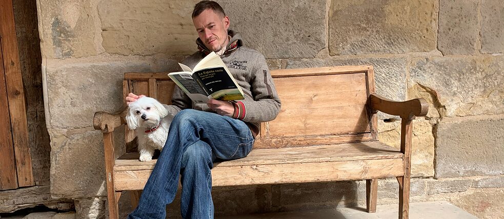 Le rédacteur web Johannes von Stritzky lit « L'Espagne vide » de Sergio del Molino sur un banc à La Fresneda, accompagné de la chienne maltaise Kika.