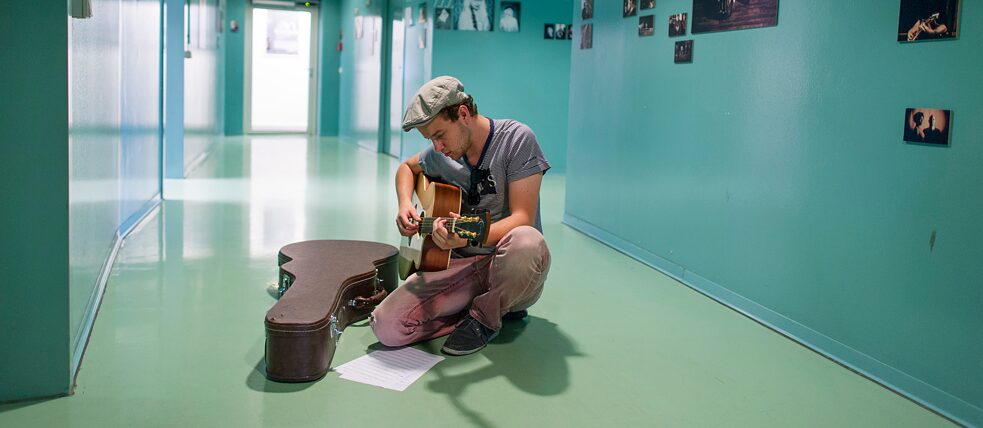 受到音樂繆思眷顧的人，流行音樂學院所有開給有志成為職業音樂家的人進修的課程，他都可以上：一位大學生在流行音樂學院的走廊上彈吉他。