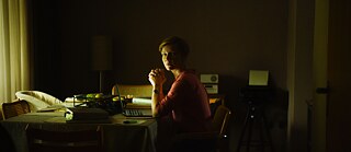 Eine Frau sitzt am Schreibtisch in einem dunklen Raum, vor ihr auf dem Schreibtisch steht ein offener Laptop