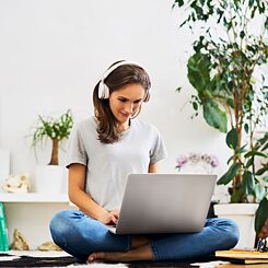 Moteris su ausinėmis sėdi namuose ant grindų ir dirba su nešiojamu kompiuteriu. Šalia matosi kambariniai augalai.