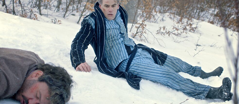Ein Mann im Morgenmantel und Schlafanzug sitzt im Schnee. Neben ihm liegt ein Mann mit Wunde, sein Gesicht im Schnee.