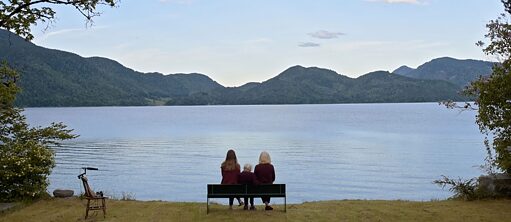 Kolm inimest istuvad järve kaldal pingil. Taamal mäed.