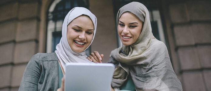 Zwei junge Frauen sitzen vor einem Gebäude und schauen gemeinsam auf ein Tablet. Sie lachen und haben Spaß