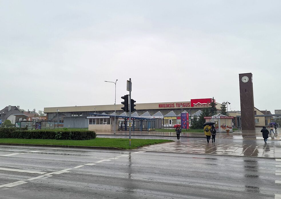 eine straße mit Ampel und Zebrastreifen, vier Leute überqueren die Straße, weiter sieht man ein großes Gebäude mit den Überschrift auf Litauisch „Neuer Markt“