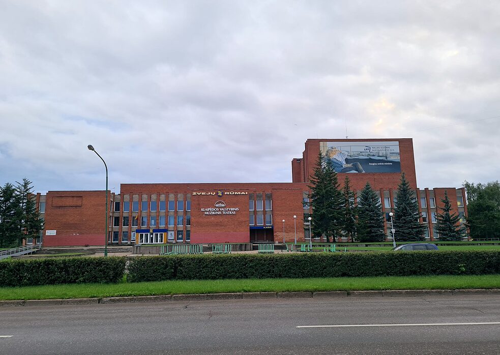 Raudonų plytų pastatas, ant fasado užrašai „Žvejų rūmai“ ir „Klaipėdos muzikinis valstybinis teatras“, priekyje gatvė su žalia zona.