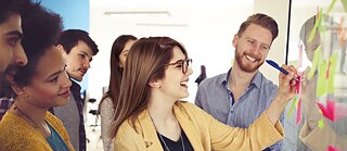 Eine Gruppe von Menschen vor einer Tafel mit mehreren Post-It-Zetteln. Auf einem Zettel schreibt eine Frau mit einem Kugelschreiber