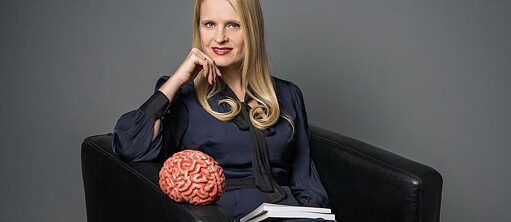 Prof. Dr. Michaela Sambanis - eine blonde Frau in einem dunkelblauen Kleid sitzt in einem schwarzen Sessel, in einer Hand hat sie Bücher, auf der Sessellehne liegt ein Gehirnmodel.
