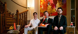 Die drei Musiker von Velvet Revolution auf einer Bank in einem Wohnzimmer sitzend. Daniel Erdmann hält in der Mitte den Hörer eines Nostalgietelefons an sein Ohr.