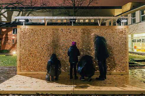 Vidiniame kieme stovi sienos instaliacija iš organinio stiklo, į kurią sutalpintos medinės kaladėlės su išgraviruotomis citatomis. Žmonės stovi ir renkasi kaladėles.