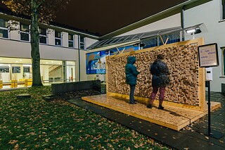 Im Innenhof steht eine Wandinstallation aus Plexiglasgerüst, in das Holzklötze mit eingravierten Zitaten eingesetzt sind. Leute wähle die Holzklötze aus.