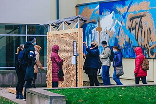 Im Innenhof steht eine Wandinstallation aus Plexiglasgerüst, in das Holzklötze mit eingravierten Zitaten eingesetzt sind. Leute wähle die Holzklötze aus.