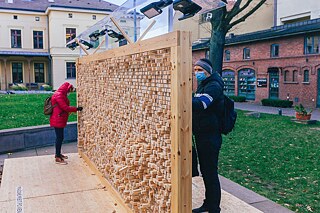 Im Innenhof steht eine Wandinstallation aus Plexiglasgerüst, in das Holzklötze mit eingravierten Zitaten eingesetzt sind. Leute wähle die Holzklötze aus.