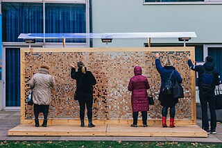 Im Innenhof steht eine Wandinstallation aus Plexiglasgerüst, in das Holzklötze mit eingravierten Zitaten eingesetzt sind. Leute wählen die Holzklötze aus.