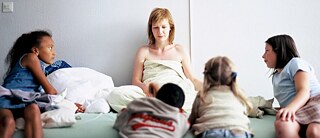 Frau sitzt beschienen von Sonnenstrahl im Bett; Kinder davor schauen sie an 