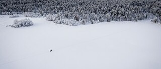 A snowy landscape from above, two people wading through the snow