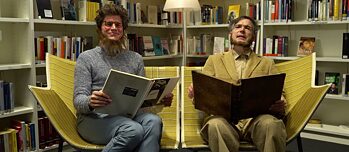 Dva muži sedí s knihami na pohovce před policí v knihovně.