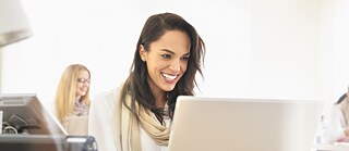 Frau mit weißer Bluse und beige Schal lächelnd am Computer. Im Hintergrund noch eine Frau am Computer unscharf.