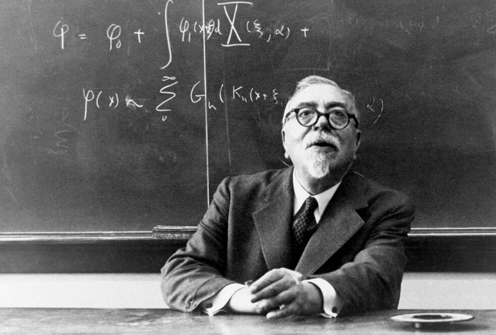 Norbert Wiener, US mathematician, sitting at a desk in front of a blackboard