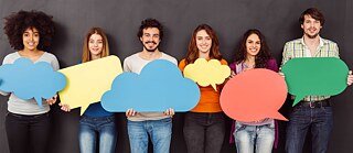 Eine Gruppe Männer und Frauen stehen nebeneinander vor einer dunklen Wand und halten große, bunte Sprechblasen vor sich. Sie haben Spaß und lachen in die Kamera