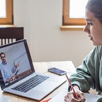 Een jonge vrouw leert Duits op haar laptop. Op het scherm is een leraar te zien. 