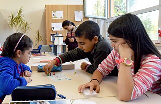 Crianças migrantes em uma aula de boas-vindas na Escola de Ensino Fundamental I Weidenhof, em Potsdam (Brandemburgo). Além das aulas regulares, crianças filhas de requerentes de asilo e refugiados recebem até cinco aulas adicionais por semana. Em 2015, cerca de 320 crianças, das quais 137 tinham histórico migratório, estavam matriculadas na escola. Há um alojamento para requerentes de asilo na área de abrangência da escola.