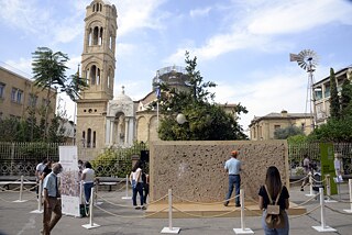 The installation at Faneromeni Square, in front of an Orthodox church, between two roll-up banners. Ten visitors are standing or walking around the wall.