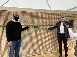 The Ambassador and the Turkish Cypriot Mayor of Nicosia are standing in front of the installation and are about to untie a green ribbon. Someone is holding a camera in front of them.