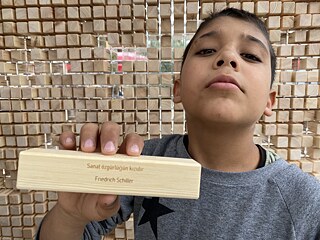 A boy in a grey sweater stands in front of the wall and shows a wooden block with a quote from Schiller engraved on it in Turkish.