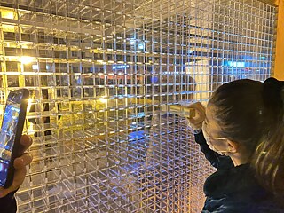 Night picture. One girl pulls a wooden block from an empty plexiglass frame. On her right we can see a hand taking a photo with a mobile phone.