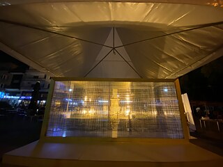 Nachtbild. Die gesamte Installation, die aus einem Holzrahmen, einem Holzsockel und einem transparenten Plexiglasrahmen besteht, befindet sich unter einem Zelt.