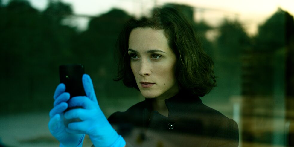 "Parfum - Ambra": Nadja Simon (Friederike Becht) is seen behind a window, looking outside. She takes a photo with her smartphone, wearing blue latex gloves.